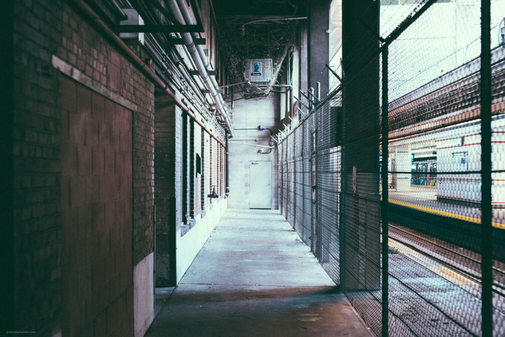 Narrow industrial alley with overhead pipes, a chain-link fence, and a door at the end