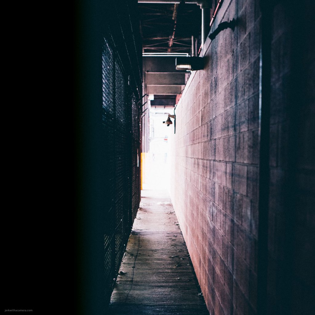A narrow, shadow-filled industrial alley lined with overhead pipes and a chain-link fence, leading to a mysterious white door at the end.