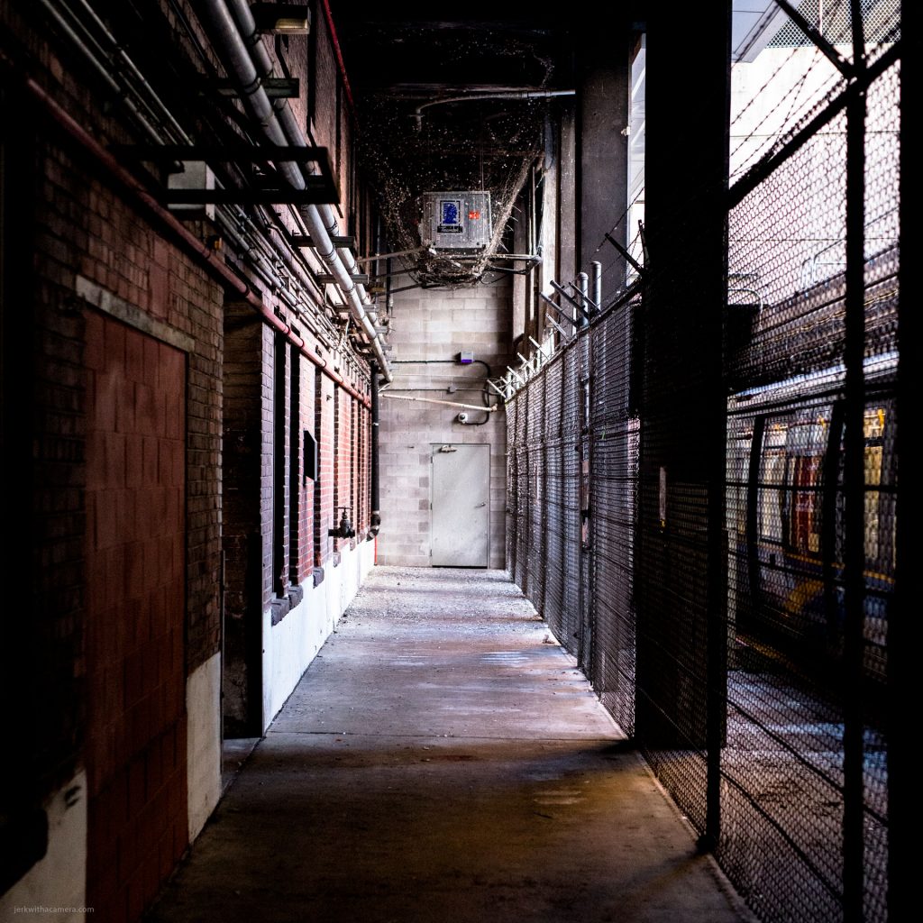 Narrow industrial alley with overhead pipes, a chain-link fence, and a door at the end