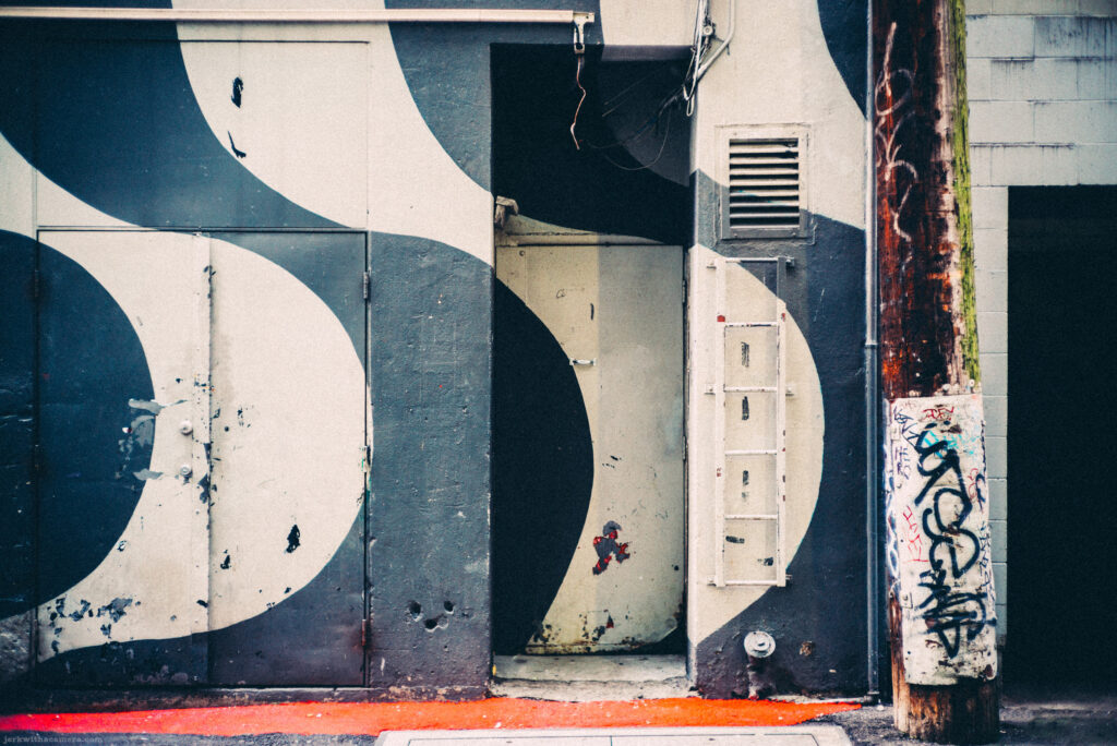 The stark contrast of graffiti against a geometric mural captures the essence of street art on Commercial Drive, Vancouver, through the sharp focus of a Leica M-D (Typ 262) paired with a Canon 50mm f/1.4 LTM."