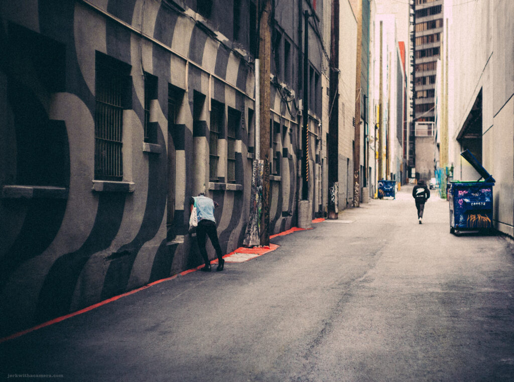 A solitary figure captured in motion down an urban alleyway, flanked by the textured walls of Emily Carr University, Vancouver, photographed with a Leica M-D (Typ 262) and Canon 50mm f/1.4 LTM lens.