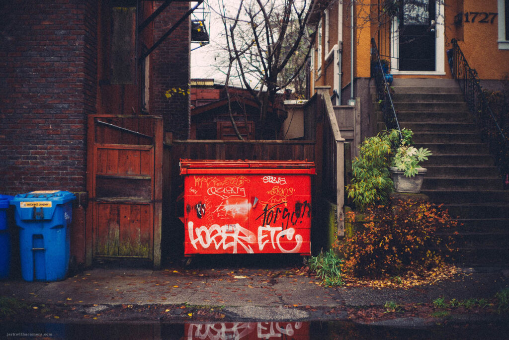 Urban alleyway on Commercial Drive in Vancouver with vibrant red graffiti on a dumpster, reflecting a scene of daily life captured with a Leica M-D (Typ 262) rangefinder and Canon 50mm f/1.4 LTM lens.