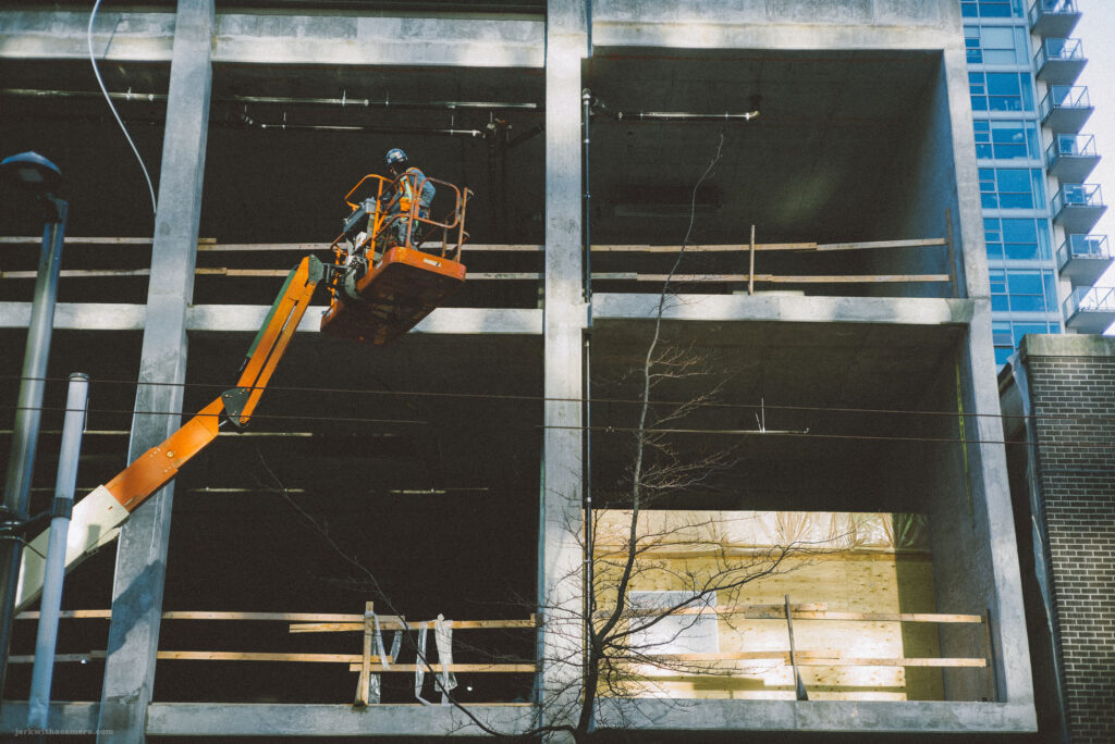"Street photography captures construction workers high on a cherry picker against the concrete skeleton of a building, shot with a Leica M-D (typ 262) Rangefinder and Canon 50mm f/1.4 LTM lens."
