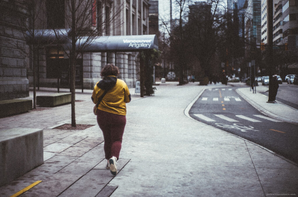 A person in a yellow jacket and maroon pants walks away from the camera on a city sidewalk, with the sign for the Vancouver Art Gallery visible in the background.