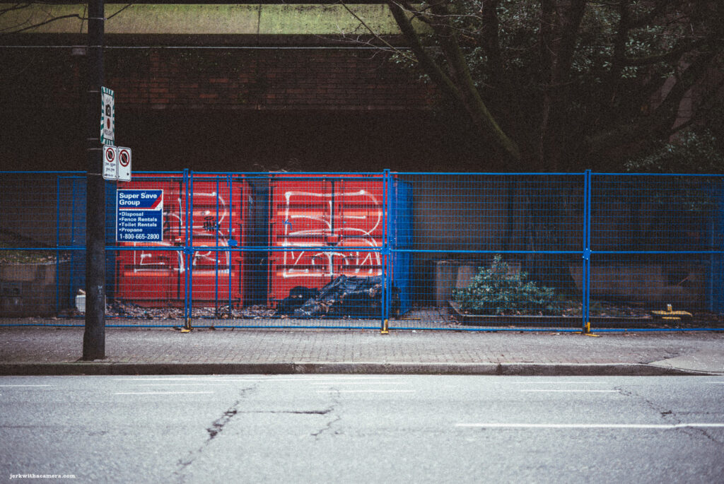 Red waste disposal containers seen through a vivid blue construction fence under an overpass in Vancouver.