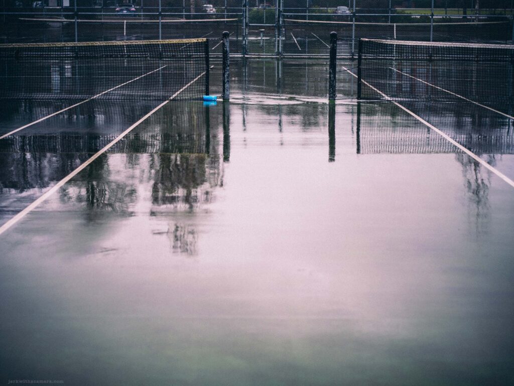 Rain-soaked tennis court with net reflecting on the wet surface, capturing the tranquility of an urban park in the rain