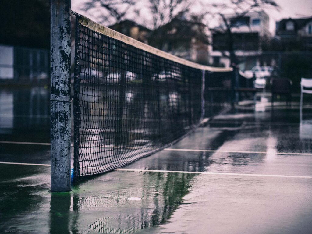 Rain-soaked tennis court with net reflecting on the wet surface, capturing the tranquility of an urban park in the rain