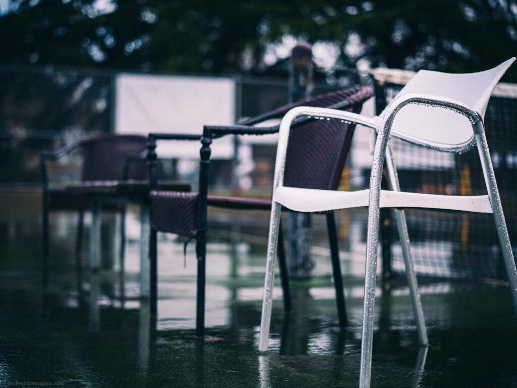 Rain-soaked tennis court with a set of plastic chairs reflecting on the wet surface, capturing the tranquility of an urban park in the rain