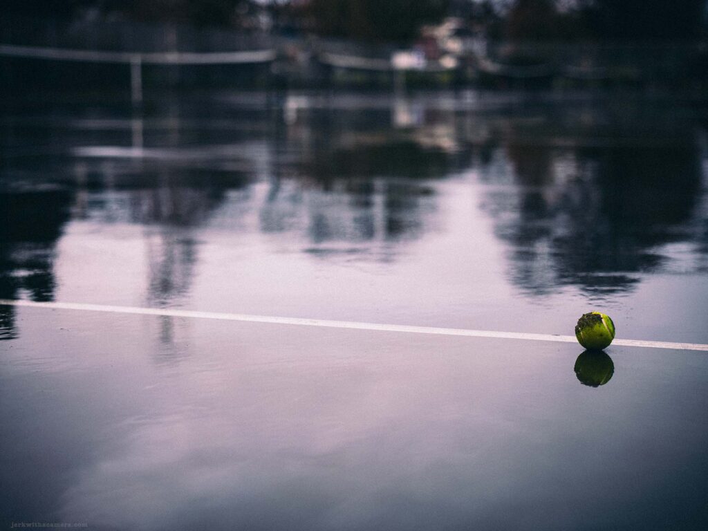 Rain-soaked tennis court with a tennis ball reflecting on the wet surface, capturing the tranquility of an urban park in the rain