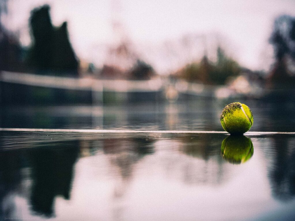 Rain-soaked tennis court with a tennis ball reflecting on the wet surface, capturing the tranquility of an urban park in the rain