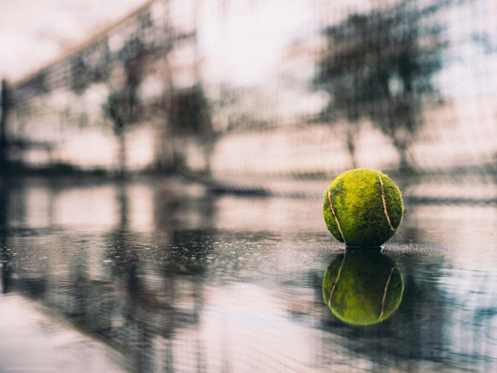Rain-soaked tennis court with a tennis ball reflecting on the wet surface, capturing the tranquility of an urban park in the rain