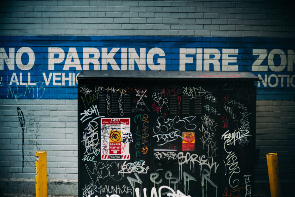A graffiti-covered utility box against a brick wall with "No Parking Fire Zone" signage.