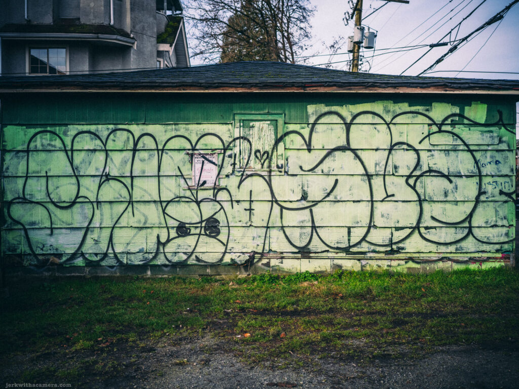 A weathered garage painted green, covered in graffiti, with peeling paint and a gritty urban backdrop.