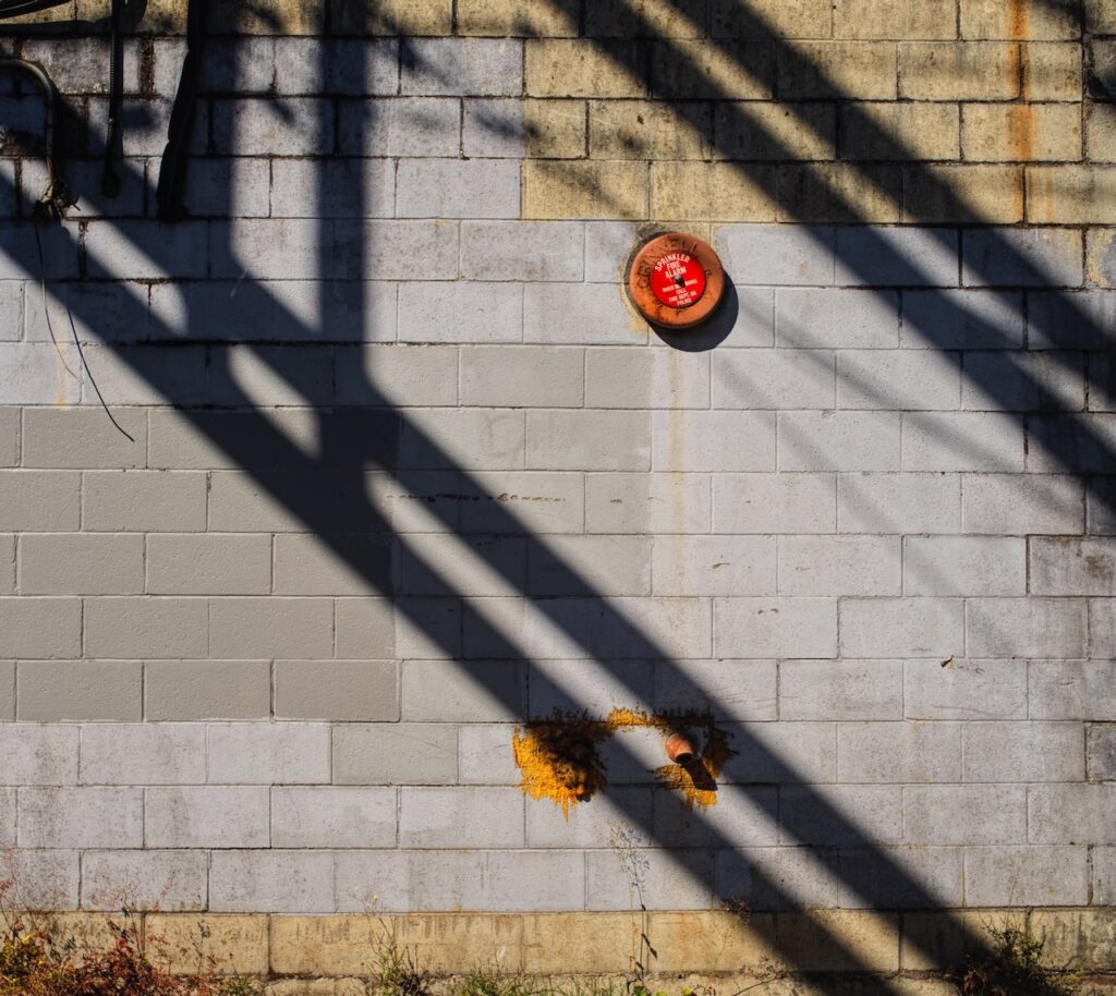 Concrete block wall with pipes, a red fire alarm bell, yellow rust stain, and shadows.