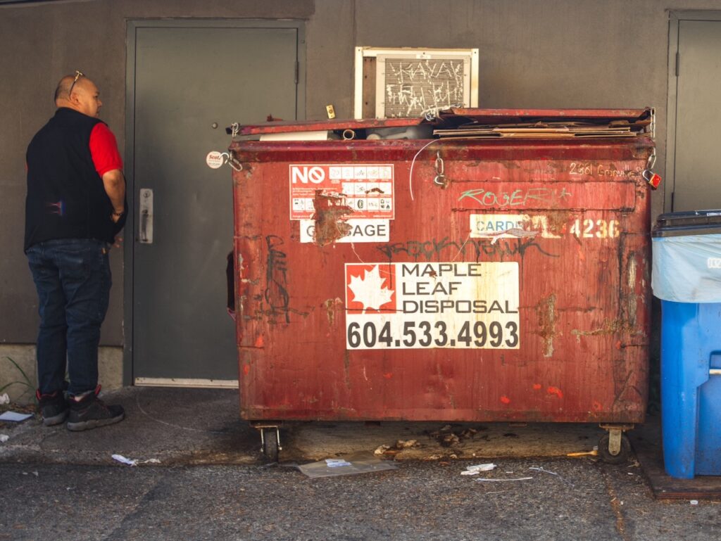 A man standing by a door next to a red dumpster with graffiti.