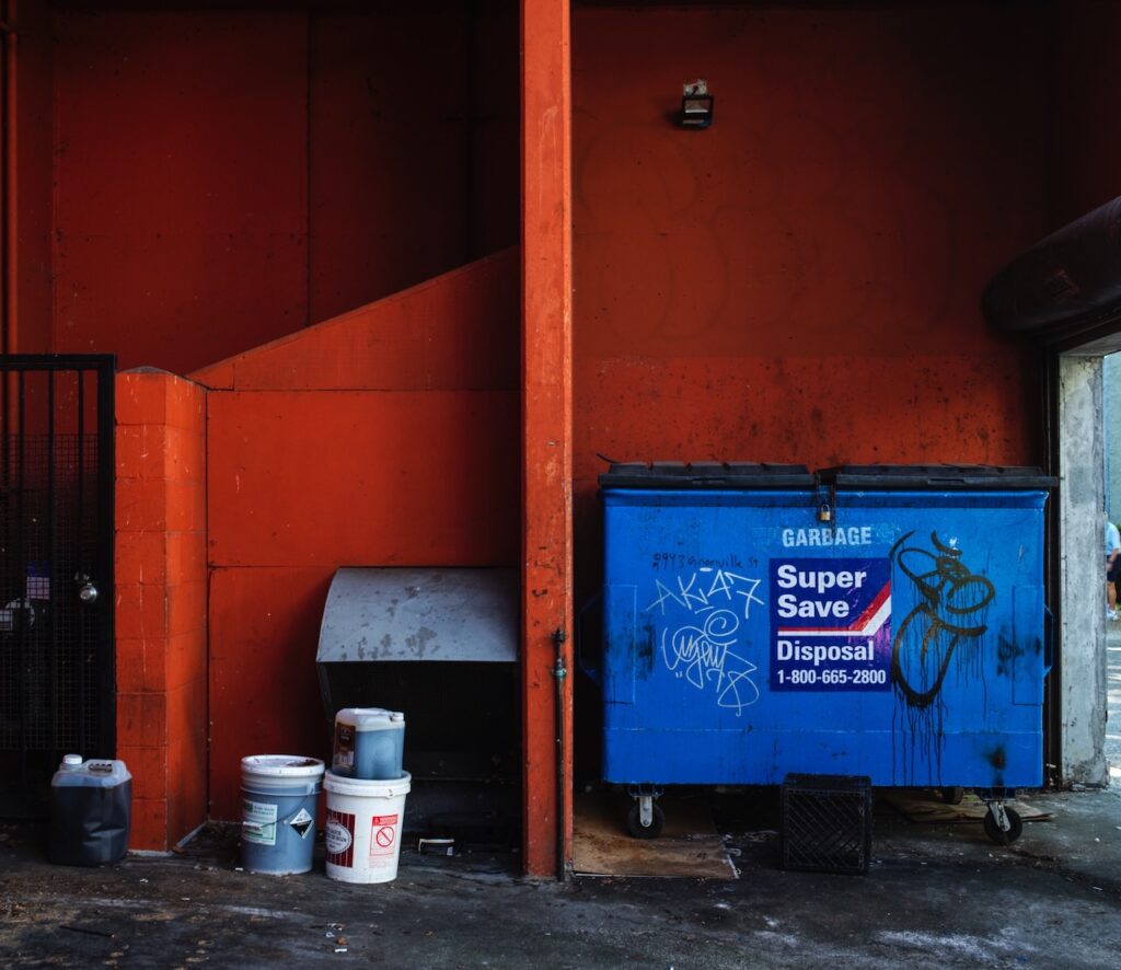 Blue dumpster against an orange wall with paint buckets nearby.