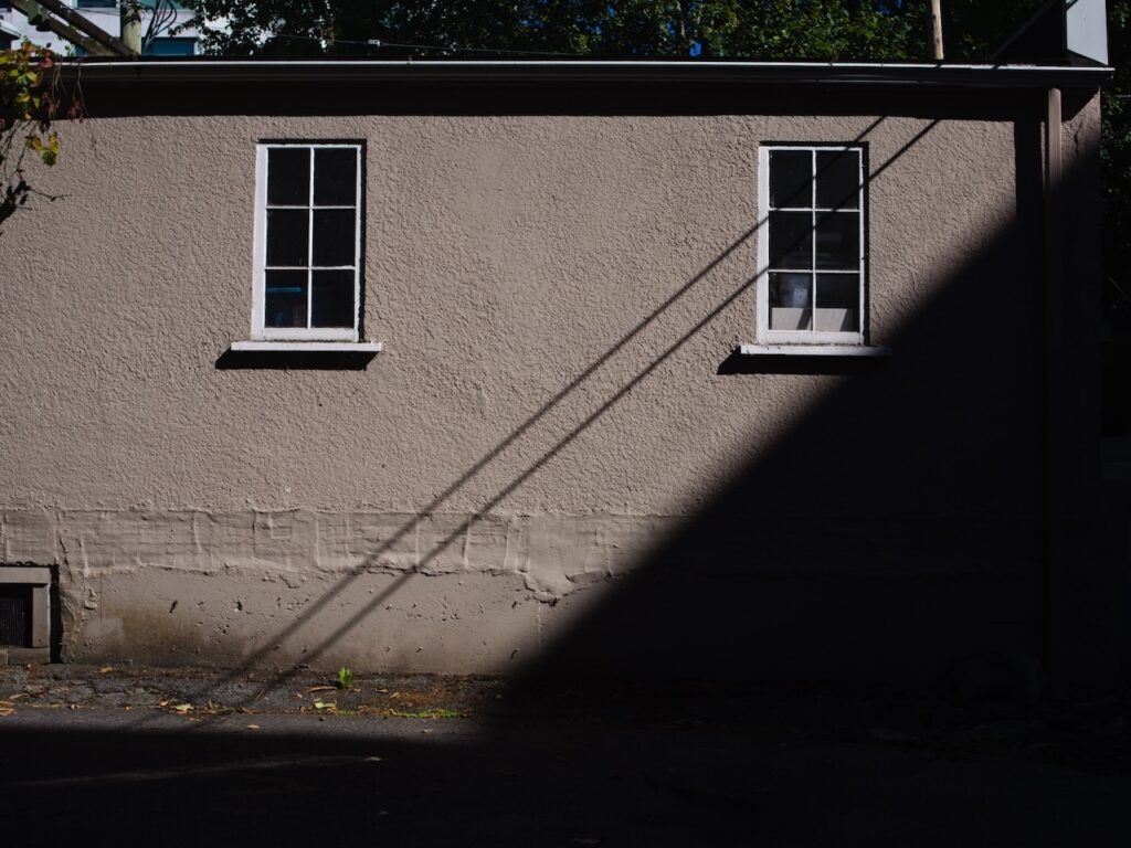 Two windows on a beige stucco wall with a diagonal shadow.