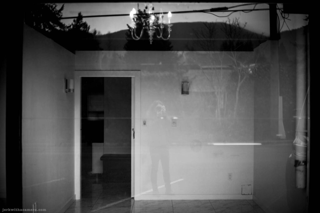 Reflection of a chandelier and a person holding a camera in a storefront window with mountains and trees in the background